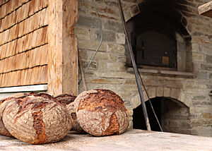 Neben einem Holzbackofen liegen zwei kräftig ausgebackene, rustikal aufgerissene Brotlaibe.