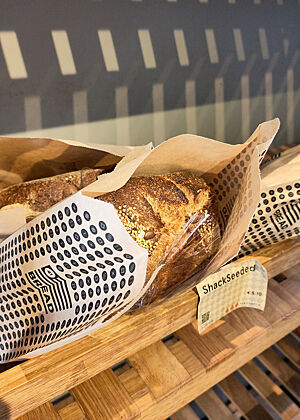 Ein Sauerteig-Saatenbrot liegt verpackt in einem Holzregal.