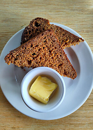 Zu zwei Dreiecken halbierte Scheibe Rübensirup-Brot liegt mit etwas Butter auf einem Porzellanteller.