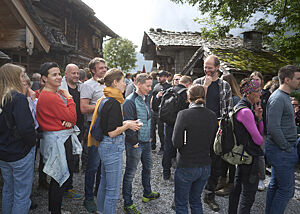 Im Sonnenschein stehen viele Menschen zwischen der Almhütte und den Holzbacköfen im Gespräch beieinander.