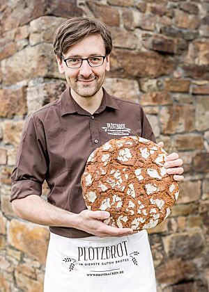 Vor einer historischen Steinmauer hält Lutz Geißler stolz ein rundes Roggenbrot mit rustikal aufgerissener Kruste in die Kamera.