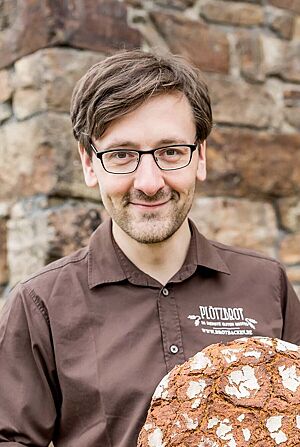 Vor einer historischen Steinmauer hält Lutz Geißler stolz ein rundes Roggenbrot mit rustikal aufgerissener Kruste in die Kamera.