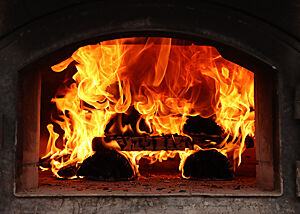 Durch die Öffnung des Holzbackofens sind brennende Holzscheite und Flammen zu sehen.