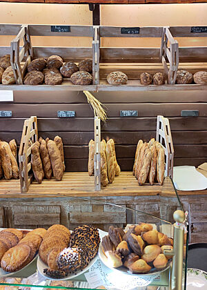 Eine Backwarenauslage in Holzoptik mit vielen Baguettes und Broten im Hintergrund. Im Vordergrund werden Madeleines und anderes Süßgebäck präsentiert.