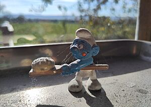 Eine Schlumpffigur mit einem Brotschieber und einem Brot darauf in den Händen steht auf einer Metallschale in der Fensterbank, durch die die Sonne ins Backhaus scheint.