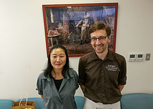 Lutz Geißler und Tomoko stehen vor einem gerahmten Bild mit Holzofenmotiv.