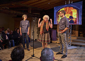 Auf einer Bühne steht Roswitha Huber – links von ihr Christina Weiß, rechts von ihr Lutz Geißler.