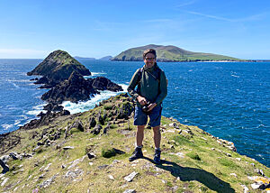 Lutz Geißler steht in Wander-Outfit mit Kamera auf den Sandsteinen des Dunmore Head, umgeben von azurblauem Wasser.