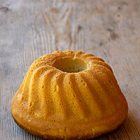 Der goldbraun ausgebackene, aus der Form gestürzte Gugelhupf liegt auf einem rustikalen Holztisch. 