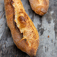 Zwei kräftig ausgebackene Baguettes, rustikal aufgebrochen auf einer rustikalen Holzunterlage.