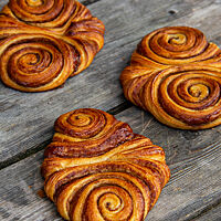 Drei goldbraune, flache Franzbrötchen mit schöner Schneckenform und glänzender Zimt-Zucker-Füllung liegen auf einem grauen Holztisch.