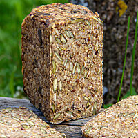 Ein kleines Kastenbrot liegt in der freien Natur auf einem Holztisch. Die Krume ist sehr dicht und besteht hauptsächlich aus Saaten und Körnern. 