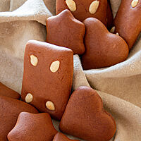 Kräftig braune Sterne, Herzen und Rechtecke mit glatt brauner, weicher Kruste und schlichter Mandelverzierung liegen auf einem cremefarbenen Leinentuch.