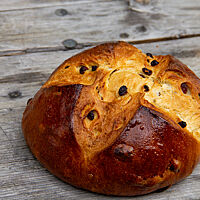 Das dunkelbraun ausgebackene Rosinenbrot mit kreuzförmigem Einschnitt auf der Oberseite liegt auf einem rustikalen Holztisch.