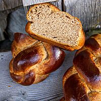 Ein dunkel ausgebackener Dinkel-Honig-Zopf mit glänzender Kruste liegt halbiert auf einem grauen Holzuntergrund.