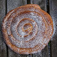 Eine goldbraun ausgebackene, mit Puderzucker bestreute Ensaïmada liegt auf einem dunkel Holztisch.