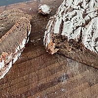 Auf einem Holzbrett liegt ein angeschnittenes, flaches Roggenbrot dessen Krume klitschig und ohne Porung ist. 