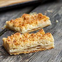 Zwei Stück Prasselkuchen liegen auf einem verwitterten Holzuntergrund. Im Anschnitt des Prasselkuchens sind im unteren Teil die Blätterteigschichten und im oberen Teil die goldbraun ausgebackenen Streusel zu erkennen.