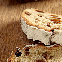 Zwei Scheiben Stollen mit vielen Rosinen in der Krume und Puderzucker auf der Kruste liegen auf einem Holztisch.