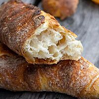 Ein durchgebrochenes, kräftig ausgebackenes Baguette mit grober, unregelmäßiger Porung liegt schräg auf einem weiteren Baguette. Im Hintergrund liegt ein weiteres Baguette längs halbiert auf einem verwitterten Holztisch. 