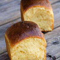 Zwei würfelförmige Brioche liegen auf einem Holztisch und zeigen die glänzende, glatte Kruste und die wattige hellgelbe Krume.