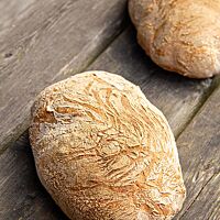 Zwei goldgelb ausgebackene Laibe Ciabatta mit glatter, leicht bemehlter Kruste liegen auf einem grauen Holztisch.