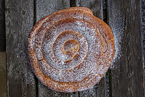 Eine große, hellbraun ausgebackene Schnecke mit Puderzucker auf der Oberfläche liegt auf einem rustikalen Holztisch.