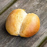 Ein goldgelb ausgebackenes Milchbrötchen mit mittig aufgerissener, glänzender Kruste liegt auf einem rustikalen Holztisch.