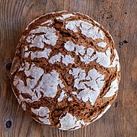 Das kräftig ausgebackene Roggenvollkornbrot mit bemehlter Kruste liegt auf einem Holztisch.