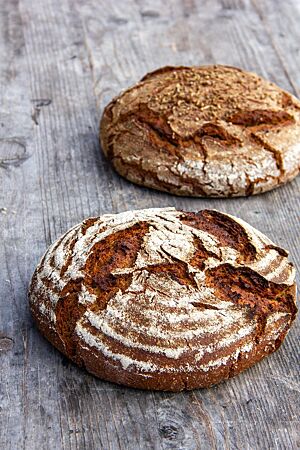 Zwei rustikal aufgerissene und kräftig ausgebackene Roggenbrote liegen auf einem Holztisch. Das vordere ist leicht bemehlt, das hintere hat Brotgewürz auf der Kruste.
