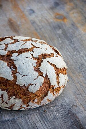 Ein kräftig ausgebackenes, leicht bemehltes Roggenbrot mit Rissen auf der Oberfläche liegt auf einem rustikalen Holztisch.