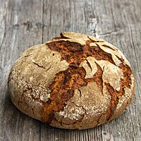 Das kräftig ausgebackene, rustikal aufgerissene Roggendinkelbrot mit leicht bemehlter Kruste liegt auf einem Holztisch.