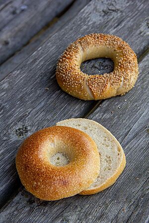 Zwei goldgelb ausgebackene Bagels liegen auf einem rustikalen Holztisch. Der hintere ist mit Sesam bestreut, der vordere hat eine glatte, leicht glänzende Oberfläche.