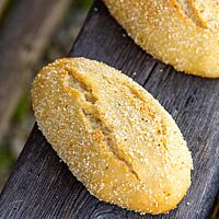 Goldgelb ausgebackene, rustikal aufgerissene Maisbrötchen liegen auf einer Holzlatte.