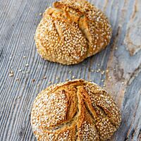 Zwei goldbraun ausgebackene, rustikal aufgerissene Weizenbrötchen mit Sesam auf der Kruste liegen auf einem Holztisch.