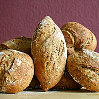 Die knusprigen, dunkelbraun ausgebackenen Brötchen liegen mit leicht bemehlter Kruste in verschiedenen Formen aufgearbeitet auf einem Tisch.