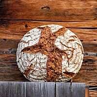 Ein kräftig ausgebackenes, rustikal aufgerissenes Vollkornbrot mit bemehlter Kruste lehnt an einer Holzwand.