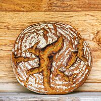 Ein runder, rustikal aufgerissener Laib mit bemehlter Kruste lehnt senkrecht an einer Holzwand.