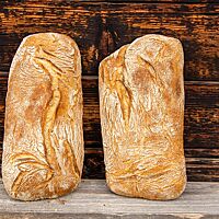 Zwei goldgelb ausgebackene Ciabattas mit leicht bemehlter und etwas aufgerissener Kruste lehnen an einer Holzwand.