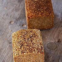 Zwei kräftig ausgebackene Kastenbrote, eines davon mit Schrot auf der Kruste, liegen auf einem grauen Holztisch.