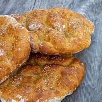 Drei runde, goldbraun ausgebackene Fladenbrote mit hellem Sesam auf der Kruste liegen gestapelt auf einem Holztisch.