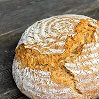 Ein runder, goldgelb ausgebackener Laib mit rustikalem Ausbund und leicht bemehlter Kruste liegt auf einem dunkelgrauen Holztisch.