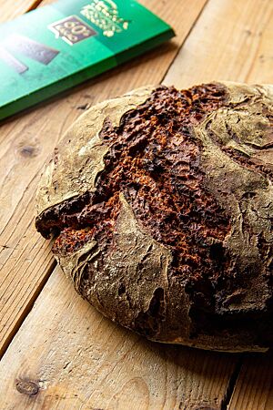 Ein kräftig ausgebackenes, rustikal aufgerissenes Roggenbrot liegt neben einer Packung Zartbitterschokolade auf einem Holztisch.