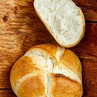 Drei goldgelbe Handsemmeln mit sternförmig aufgerissener Kruste und sehr heller, feinporiger Krume liegen auf einem Holzbrett.
