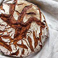 Ein kräftig ausgebackenes, rustikal aufgerissenes Roggenmischbrot mit bemehlter Kruste liegt in einem Leinentuch.
