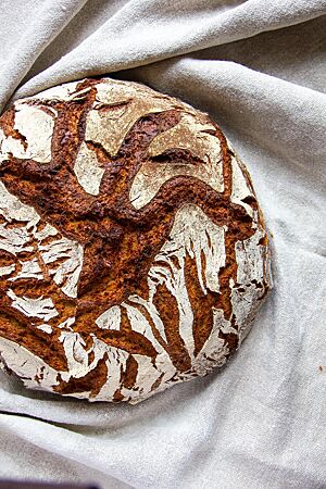 Ein kräftig ausgebackenes, rustikal aufgerissenes Roggenmischbrot mit bemehlter Kruste liegt in einem Leinentuch.