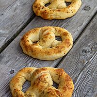 Drei dicke hellbraune Brezeln mit gemahlenem schwarzem Pfeffer auf der Kruste liegen auf einem grauen Holztisch.