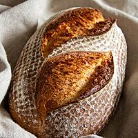 Ein goldbraun ausgebackenes, rustikal aufgerissenes Weizenmischbrot mit leicht bemehlter Kruste liegt in einem Leinentuch.
