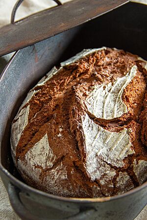 Das kräftig ausgebackene, rustikal aufgerissene Rotkornbrot mit bemehlter Kruste liegt in einem Gusseisentopf.