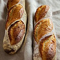 Zwei Stangenweißbrote mit dem typischen Baguette-Schnitt liegen auf einem Leinentuch.